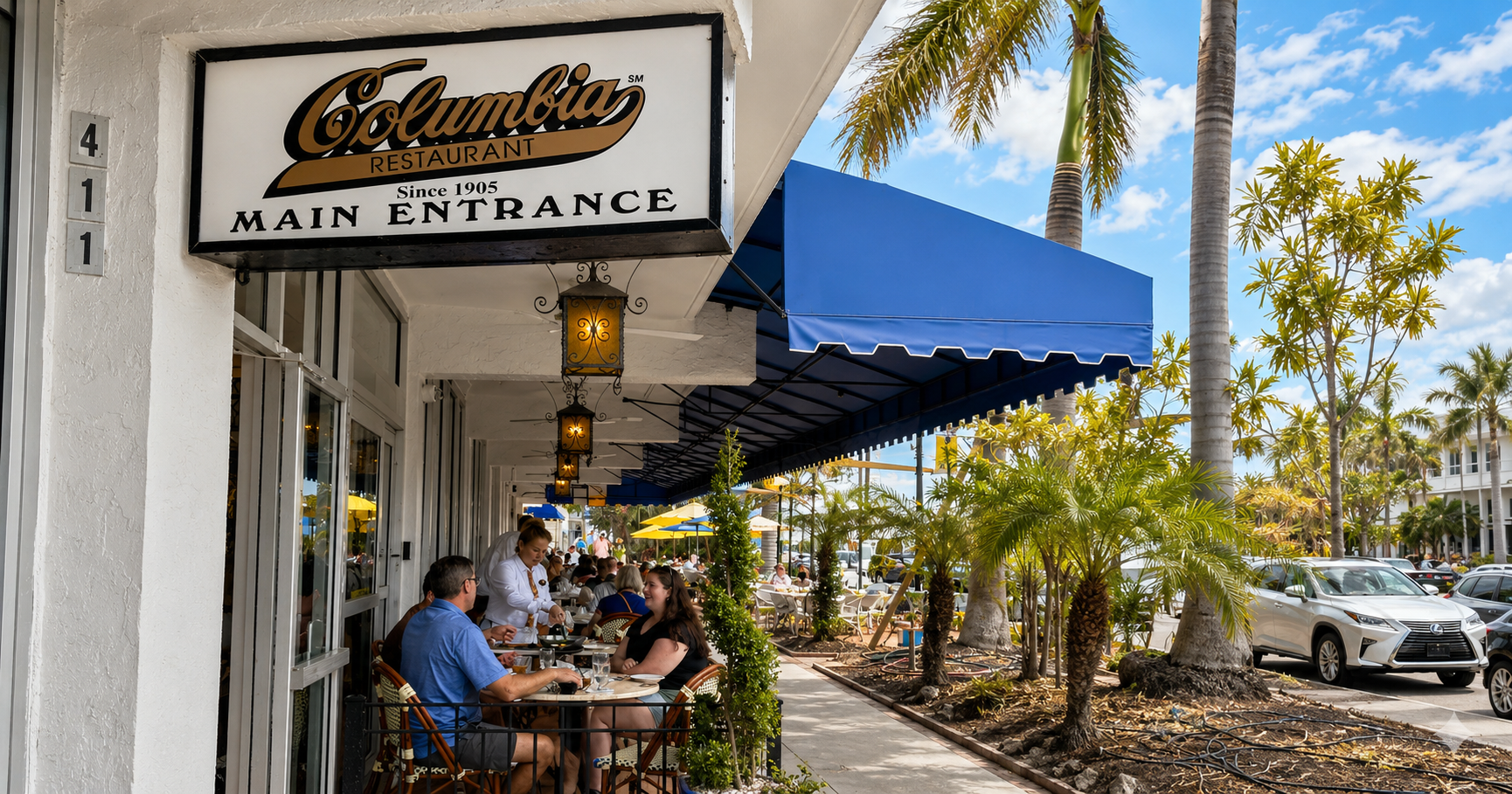 Diners seated beneath blue awnings and palm trees outside Columbia Restaurant on St. Armands Circle in Sarasota