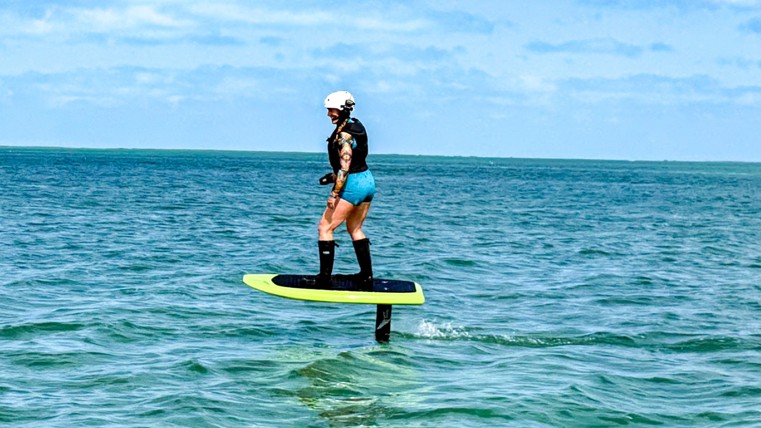 Leah Goodman wearing a helmet and life vest while gliding above blue-green water on an electric hydrofoil board
